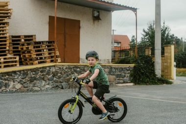 A young boy in a helmet rides his bicycle near a warehouse with stacked wooden pallets.