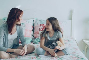 Young mother and little cheerful daughter talking and sharing secrets while sitting on bed at home in bedroom. Relationship and family concept